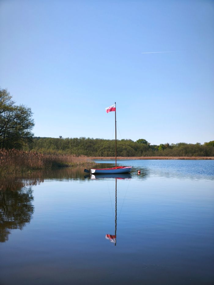Menu Tranquil scene of a sailboat with a Polish flag reflecting on a serene lake in Osieczna, Poland.